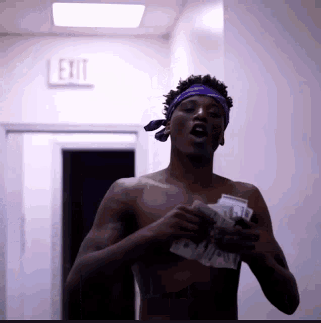 a shirtless man in a purple bandana holds a bunch of money in front of a red exit sign