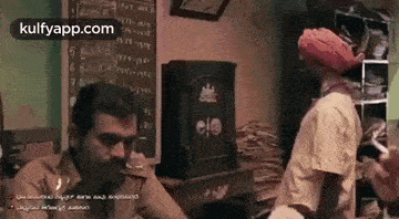 a man in a turban is talking on a cell phone while another man stands behind him .