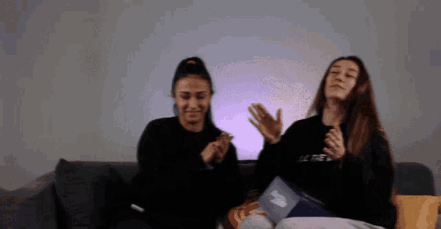 two women are sitting on a couch with one wearing a black sweater that says all the
