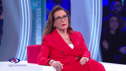 a woman in a red suit and glasses sits on a pink couch in front of a sign that says egrandellatallo