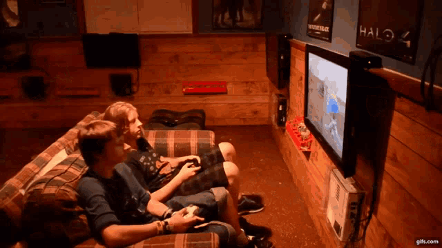 two boys sit on a couch playing a video game with a halo 4 sign on the wall behind them
