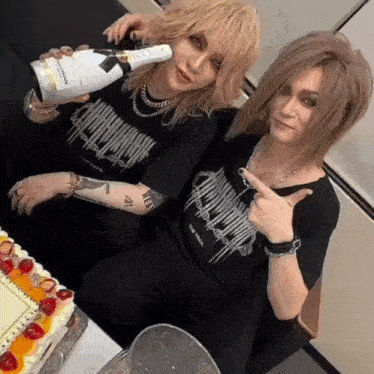 a couple of people sitting next to each other with one holding a bottle of champagne and the other holding a cake