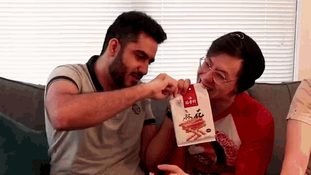 two men are sitting on a couch eating a bag of noodles .