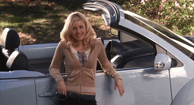a woman leans against a silver car with the top down