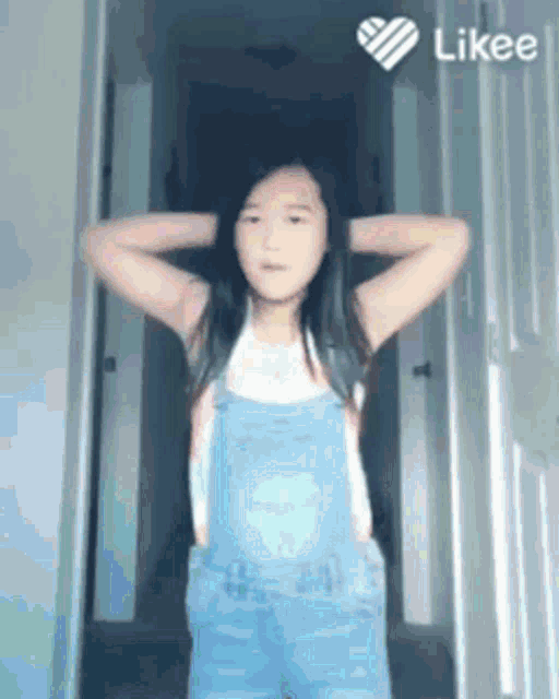 a little girl is standing in a hallway with her hands on her head and a likee heart in the corner .