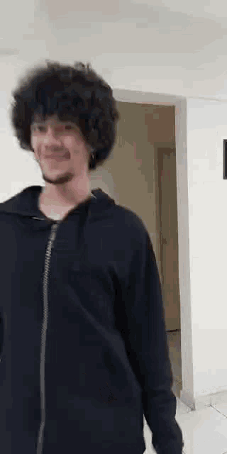 a man with curly hair and a beard wearing a black hoodie is standing in a room .