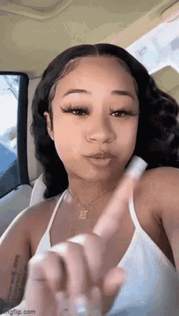 a woman in a white tank top is sitting in a car and pointing her finger .