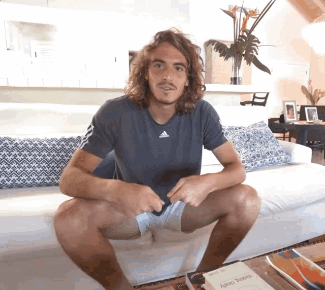 a man with long hair is sitting on a couch with his legs crossed and an adidas shirt on