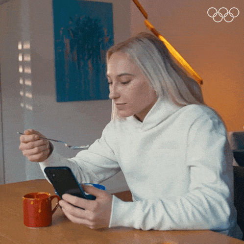 a woman sitting at a table looking at her phone and eating cereal
