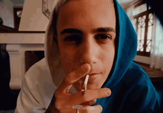 a man in a blue hoodie is smoking a cigarette with the words maharelgifs below him