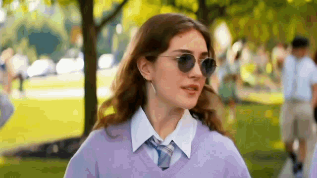 a girl wearing sunglasses and a purple sweater and tie is standing in a park .