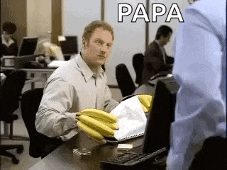 a man is sitting at a desk holding a bunch of bananas and the word papa is on the screen behind him .