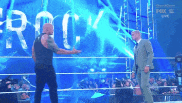 a man in a suit is standing next to a man in a black tank top in a wrestling ring .