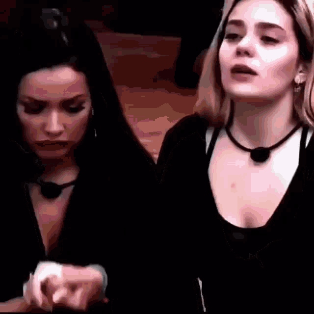 two women are standing next to each other in a dark room . one of the women is wearing a choker necklace .