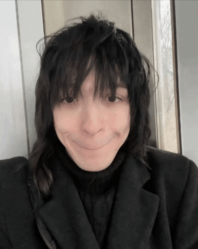 a person with long black hair and a black coat making a funny face