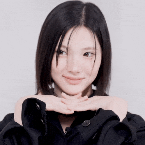 a woman in a black jacket is smiling with her hands folded under her chin