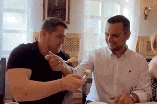 two men are sitting at a table and one is holding a spoon in his hand
