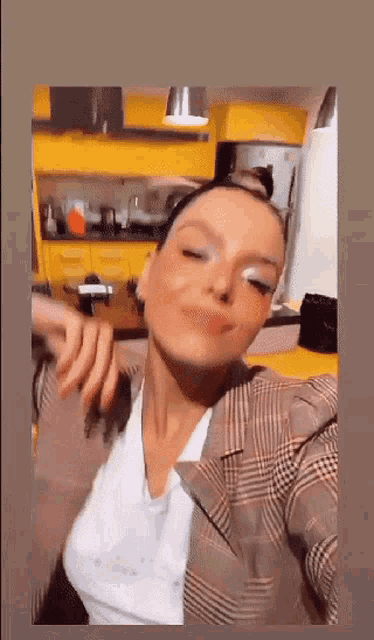 a woman is taking a selfie in a kitchen while wearing a plaid jacket and a white shirt .