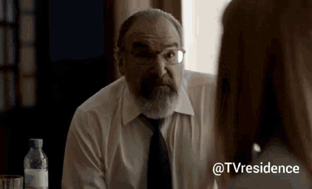 a man with glasses and a beard is talking to a woman while sitting at a table .