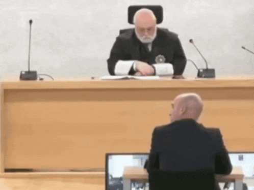 a man in a suit sits at a desk in front of a judge in a courtroom