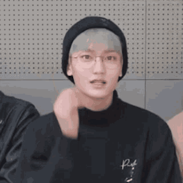 a young man wearing glasses , a beanie , and a black sweater is making a funny face .
