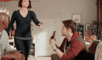 a man is holding a bottle of beer while a woman stands in the middle of a living room .