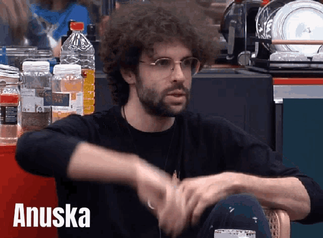 a man with curly hair and glasses is sitting in a chair with the name anuska on the bottom right