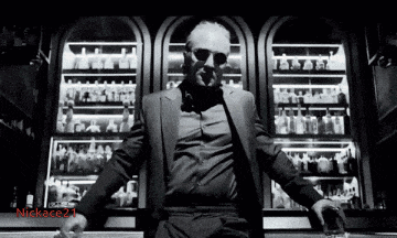 a man in a suit and sunglasses is sitting at a bar holding a glass of whiskey and smoking a cigarette