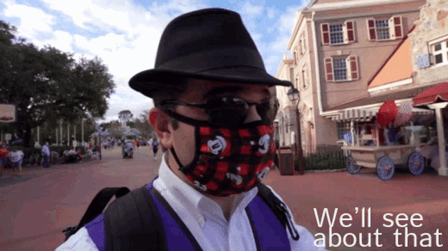 a man wearing a mickey mouse face mask stands in front of a building with the words " we 'll see about that " below him