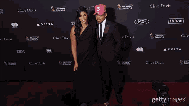 a man and a woman are standing on a red carpet in front of a clive davis wall