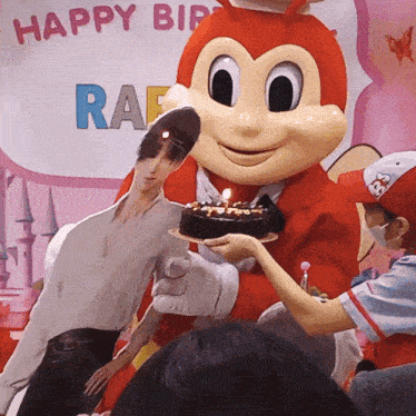 a mascot is holding a cake in front of a happy birthday sign