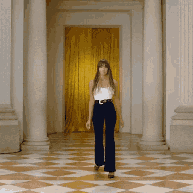 a woman in a white tank top and black pants stands in a hallway with columns