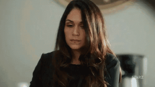 a woman in a black dress is sitting in front of a coffee pot and looking at the camera .