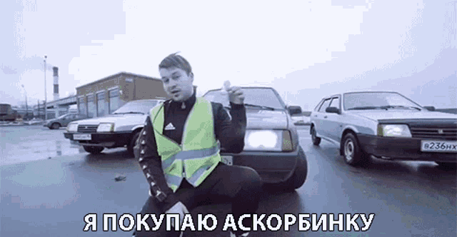 a man wearing a yellow vest is kneeling in front of a row of cars with russian writing on the bottom of the screen