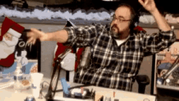 a man in a plaid shirt sitting in front of a microphone with his arms in the air