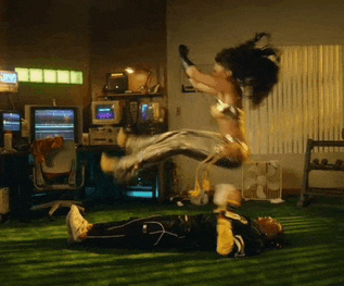 a woman in a wonder woman costume is jumping in the air over a man laying on the floor .