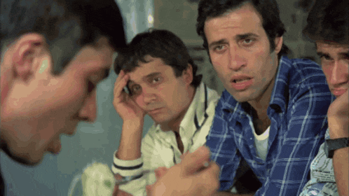 a group of men are sitting around a table and one has his hand on his forehead