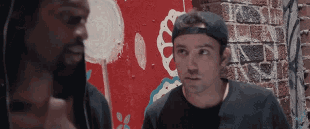 two men are standing in front of a brick wall with graffiti