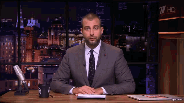a man in a suit and tie is sitting at a desk with a microphone in front of him and a sign that says 7 hd