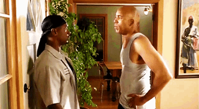 two men are standing next to each other in a living room and talking to each other .