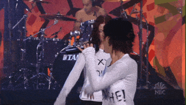 a man playing drums behind two women with one wearing a shirt that says " hate "