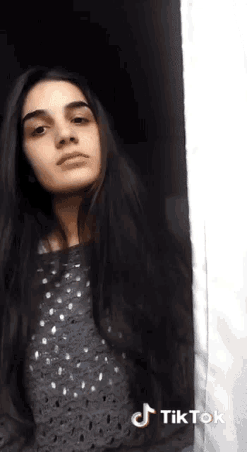a woman with long black hair is standing in front of a window with a tiktok logo on the bottom right