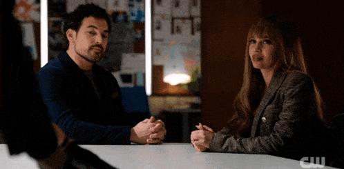 a man and a woman are sitting at a table with a cw logo on the table