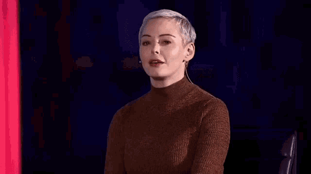 a woman with short hair is shrugging her shoulders while sitting in front of a red curtain .