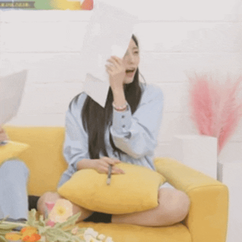 a woman is sitting on a yellow couch with a yellow pillow and covering her face with a piece of paper .
