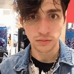 a young man wearing a denim jacket and a necklace looks at the camera