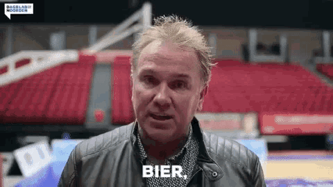 a man in a leather jacket is standing in front of a basketball court and talking .