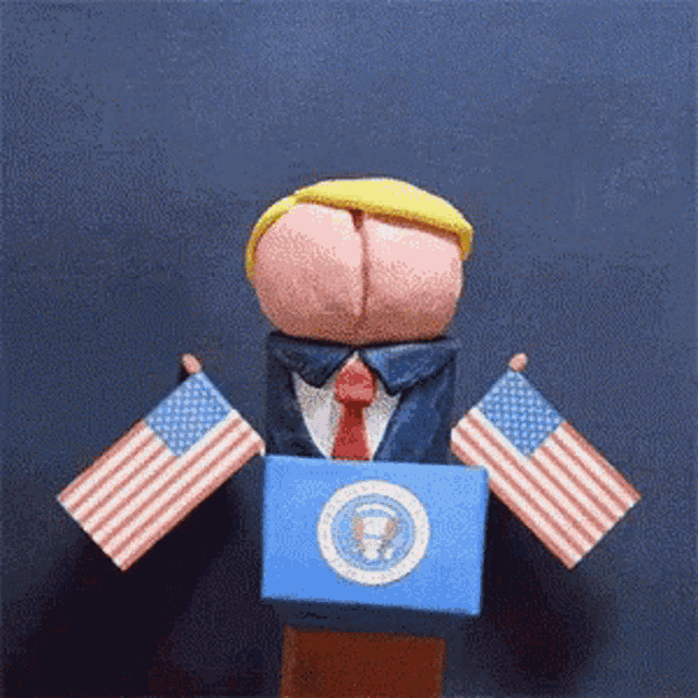 a puppet of donald trump stands behind a podium holding american flags