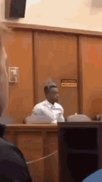 a man in a white shirt is sitting in a courtroom with a crown on his head .