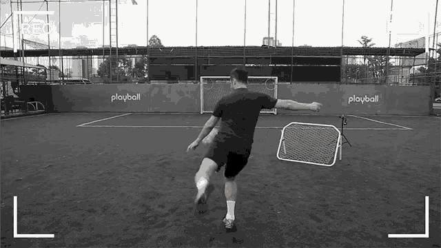 a black and white photo of a man kicking a soccer ball in front of a playball sign